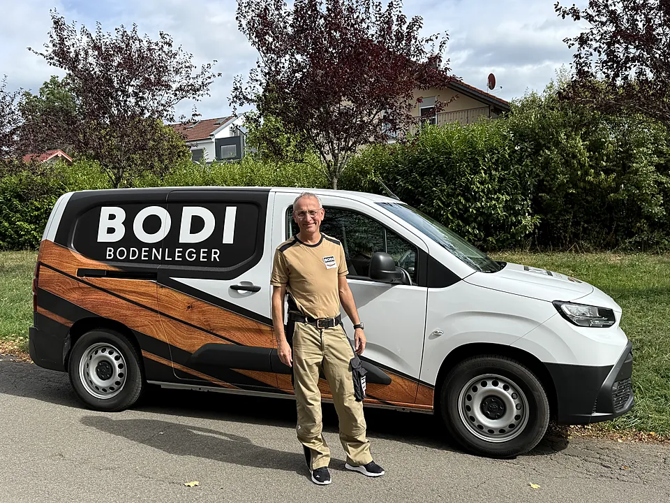 Bodenleger Bodi in Arbeitskleidung vor seinem Firmenwagen mit Logo und Holzoptik-Folierung